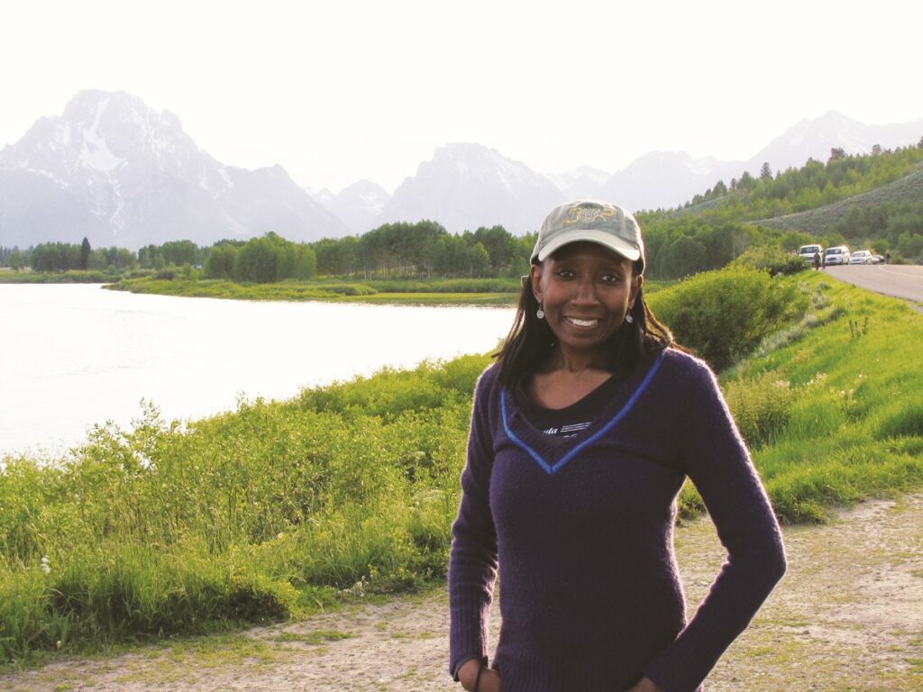 Cassandra Johnson Gaither wears a long-sleeved blue shirt and a baseball cap and stands in front of a body of water. Mountains are visible in the distance.