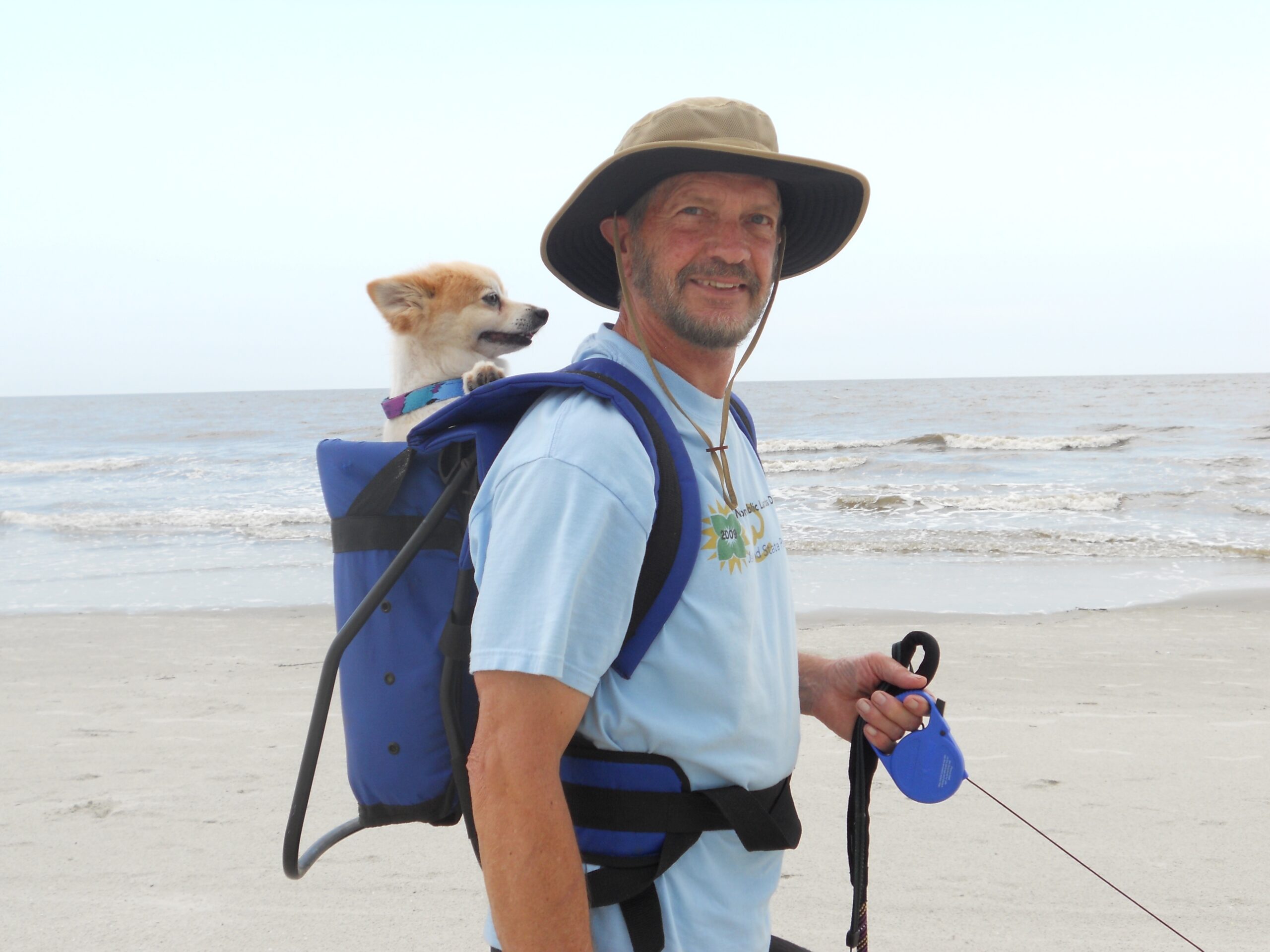 Dr. Cordell stands on a beach holding a lease with another dog in a backpack on his back.