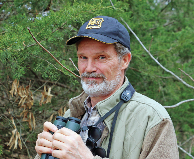 Frank Thompson stands outside wearing a baseball cap and holding a pair of binoculars.