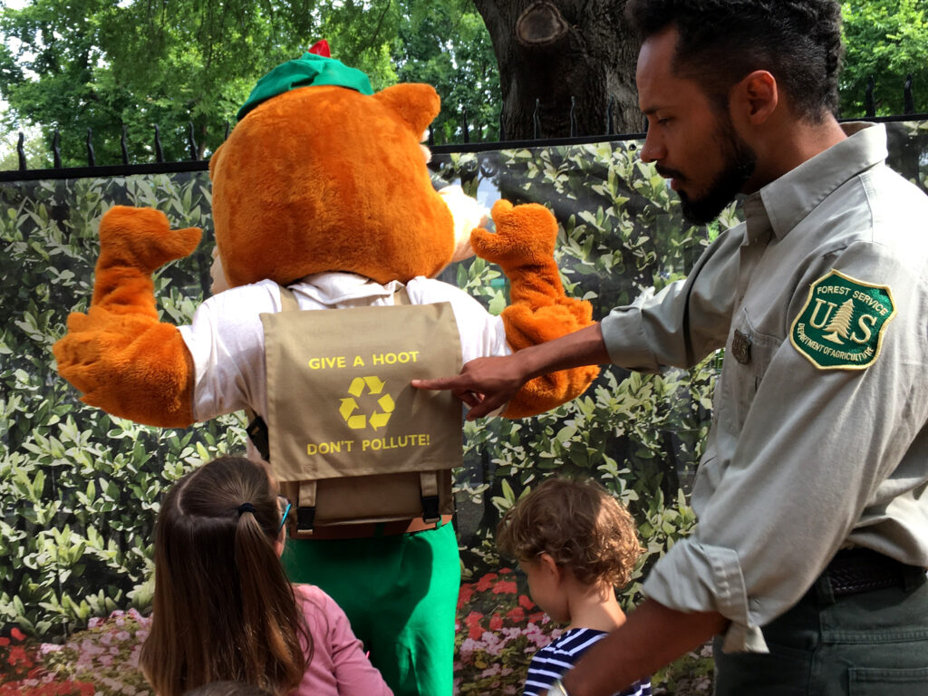 A Forest Service emplyee pointing to a reuse sign on the back on Woodsy Owl's backpack. He is showing it to a group of children.