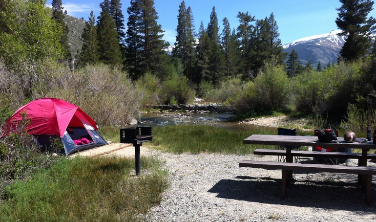 A campsite with a tent set up on the left, a small grill in the middle, and a picnic table to the right. Mountains and trees are in the background.