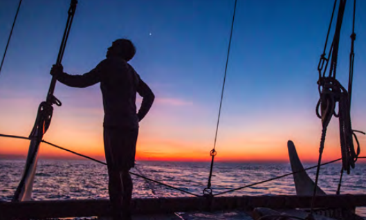 A person stands on a ship in silhouette against the setting sun.