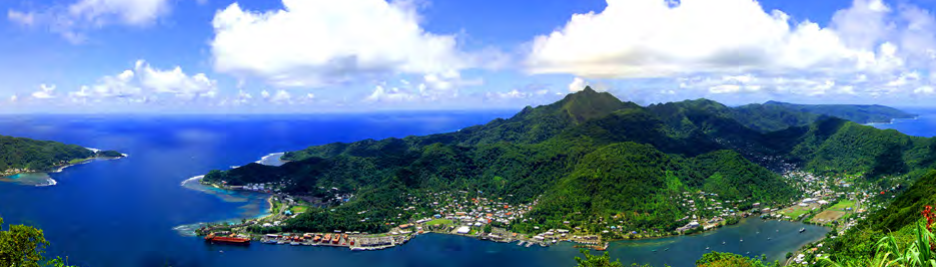 A green, mountainous island. The harbor has several buildings along the shoreline.