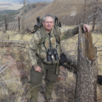 A man wearing camouflage and binoculars stands next to a tall tree stump.