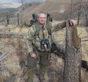 A man wearing camouflage and binoculars stands next to a tall tree stump.