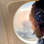 Mr. Atkins wears a headset and looks out a plane window