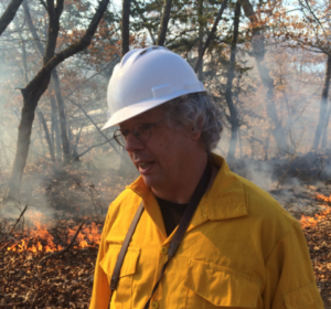 Dan Dey in protective in a forest with a small prescribed burn behind him