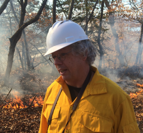 Dan Dey in protective in a forest with a small prescribed burn behind him