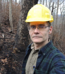 Dr. Norman wears a yellow hard hat in a forest.
