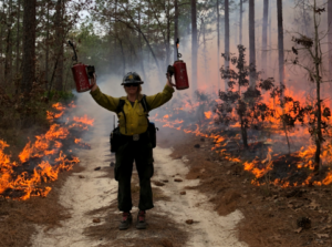 Lindsey Kupfer standing with two torches ni front of a prescirbed burn in a forest.