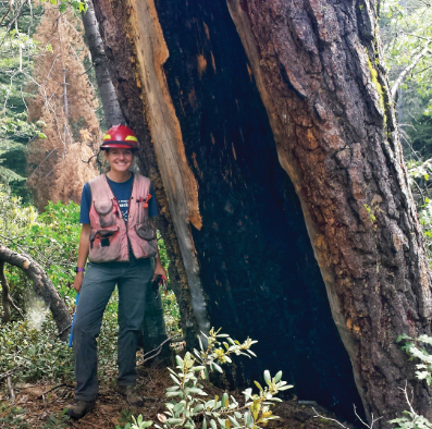 Sharon Hood in a hard hat in a forest in front of a large tree that has been burned