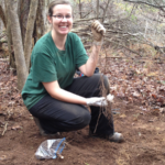 Ms. Taylor kneels on a forest floor and holds up a long plant root