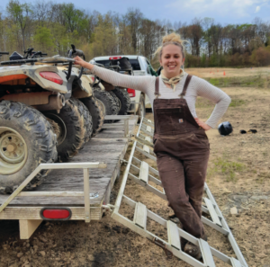 Ms. Klebanski wears overalls and stands next to a trailer loaded with ATVs.