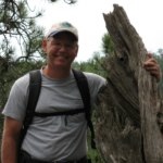 Dr. Butler wears a backpack and stands next to a tall, dead tree stump.
