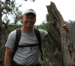 Dr. Butler wears a backpack and stands next to a tall, dead tree stump.
