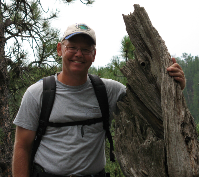 Dr. Butler wears a backpack and stands next to a tall, dead tree stump.