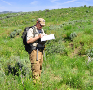 Mr. Colt takes notes on a clipboard as he stands in a grassland.